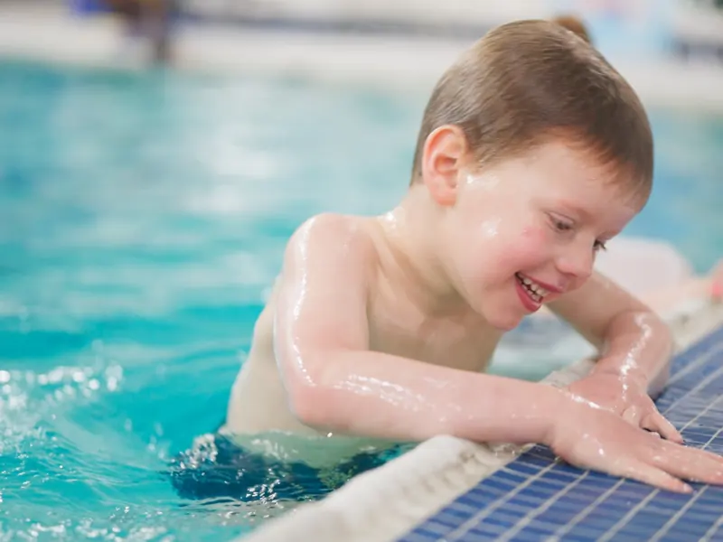 A toddler at the pool wall, grinning, water up to the shoulders during a Seal Pups lesson.