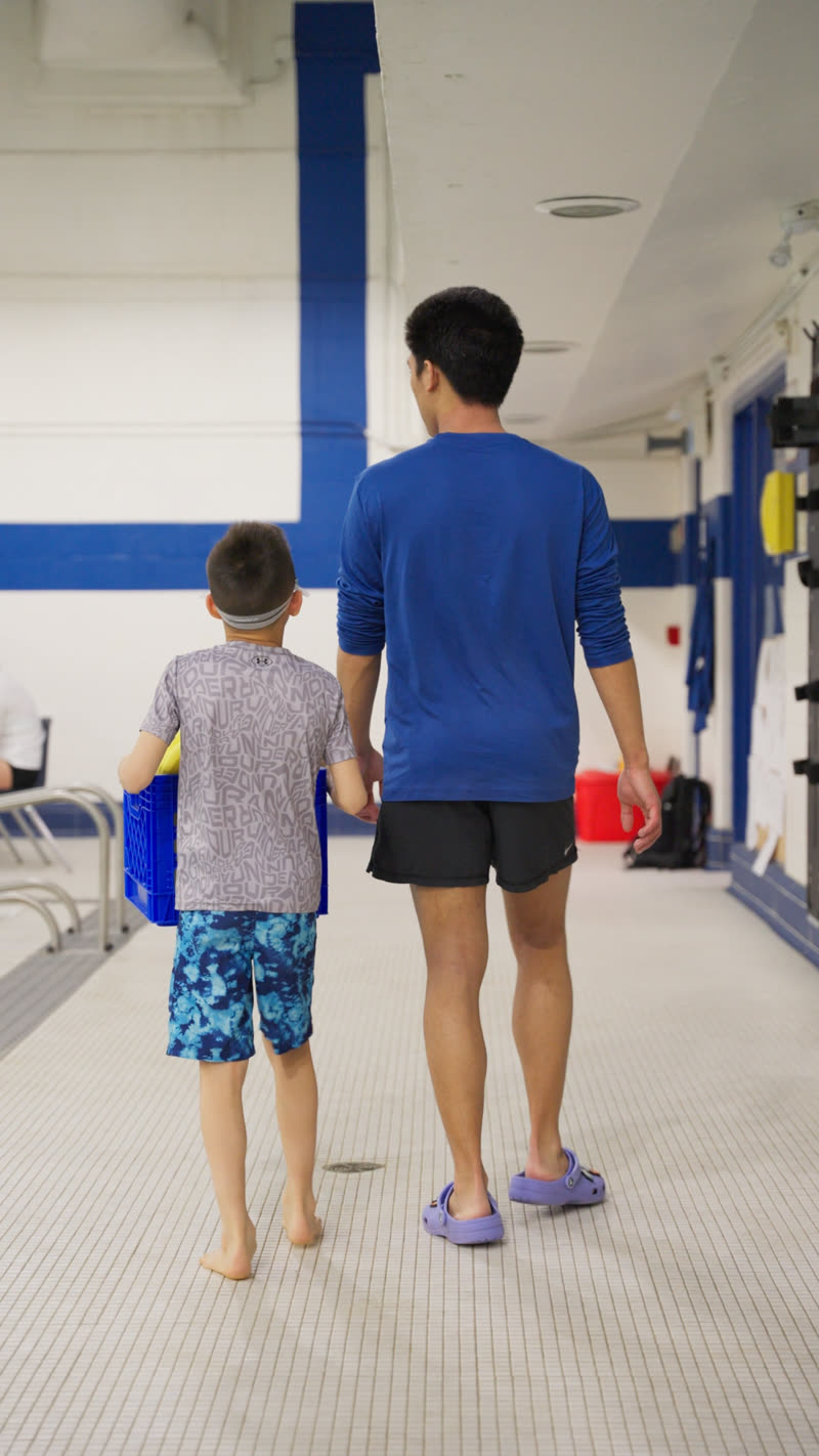 A swim teacher walks a swimmer onto the pool deck, carrying the equipment crate together.