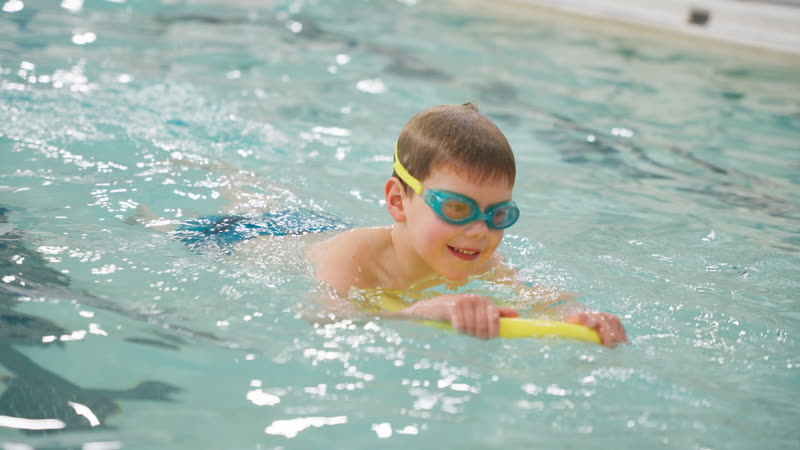 A child in goggles kicks the length of the pool holding a flutter board.