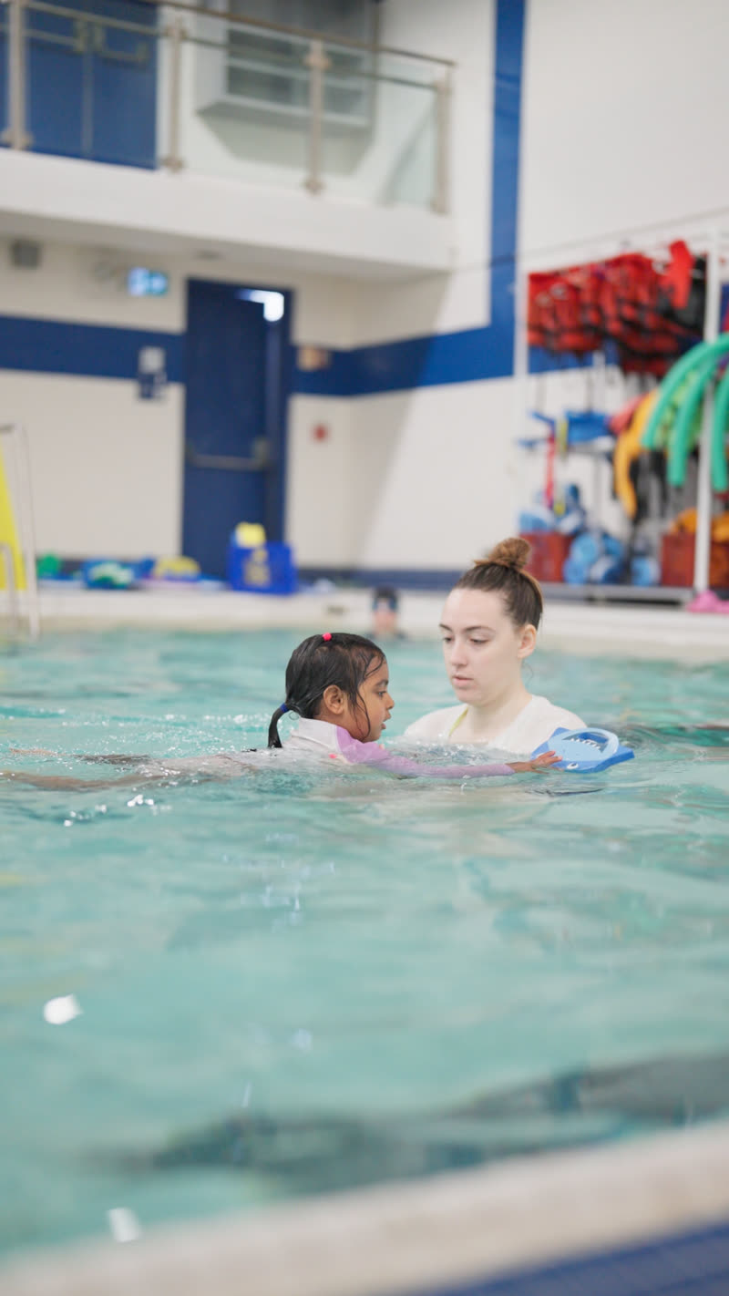 A swim teacher works one-to-one with a young swimmer using a kickboard.