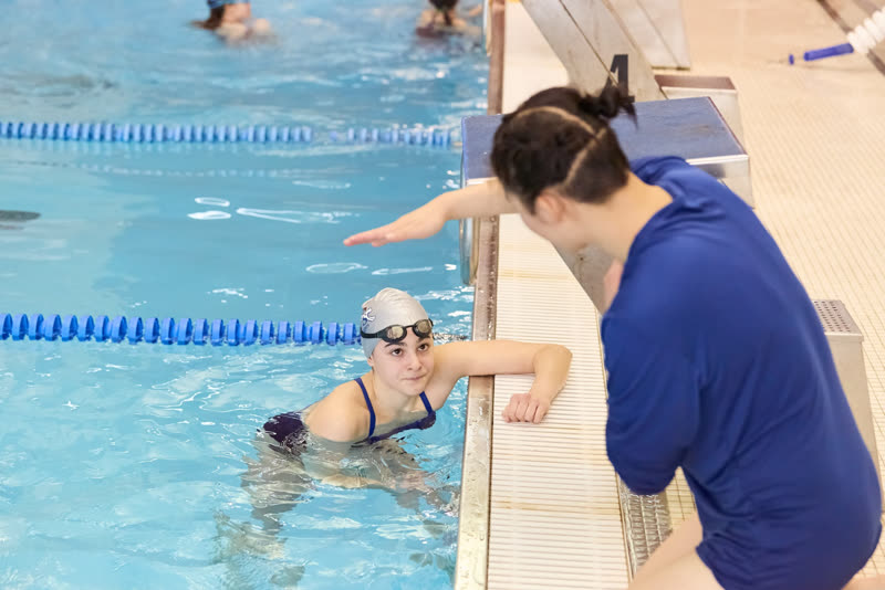 A coach on the deck gives stroke feedback to a swimmer resting at the wall.