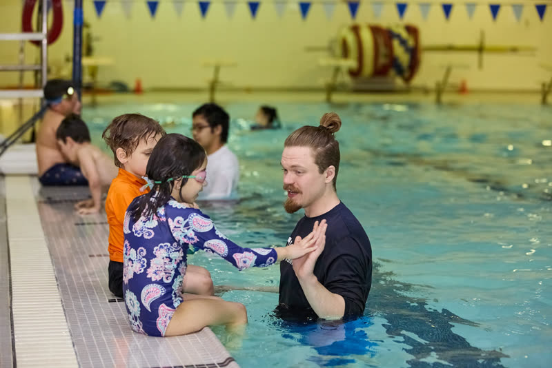 A swim teacher in the water talks with two children sitting on the pool edge.
