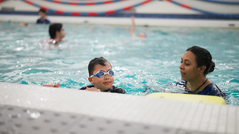 A swimmer in goggles holds the wall while their swim teacher gives feedback alongside.